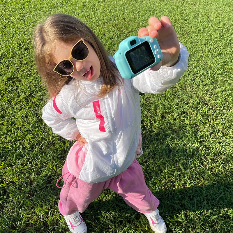 Child holding blue LittleLens Kids Camera outdoors on grass, wearing sunglasses and sporty clothes